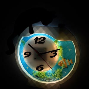 May include: A novelty clock with a fish tank design. The clock face has black numbers and hands. A cartoon clownfish and aquatic plants are visible inside the clock. The clock is illuminated with a soft glow.