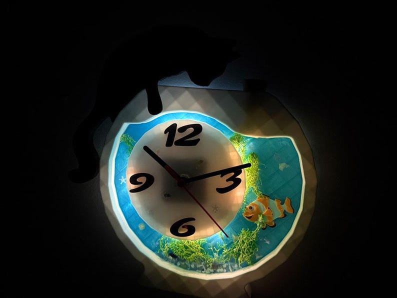 May include: A novelty clock with a fish tank design. The clock face has black numbers and hands. A cartoon clownfish and aquatic plants are visible inside the clock. The clock is illuminated with a soft glow.