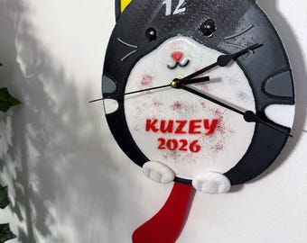 May include: A whimsical cat-shaped wall clock with a black and white design, yellow ears, and a red tongue. The clock face features the number 12 and the name "KUZEY 2026" in red. The clock hands are black.