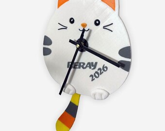 May include: A white cat-shaped wall clock with orange-tipped ears, gray spots, and a striped tail. The clock face features black hands, a small nose, and the text "PERAY 2026". A whimsical decorative item.