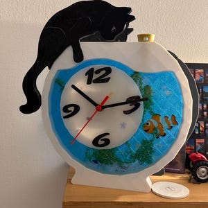 May include: A whimsical clock shaped like a fishbowl, featuring a clownfish and aquatic plants. A black cat silhouette perches atop the white frame. The clock face has black numbers and hands, with a red second hand. A unique decorative item.