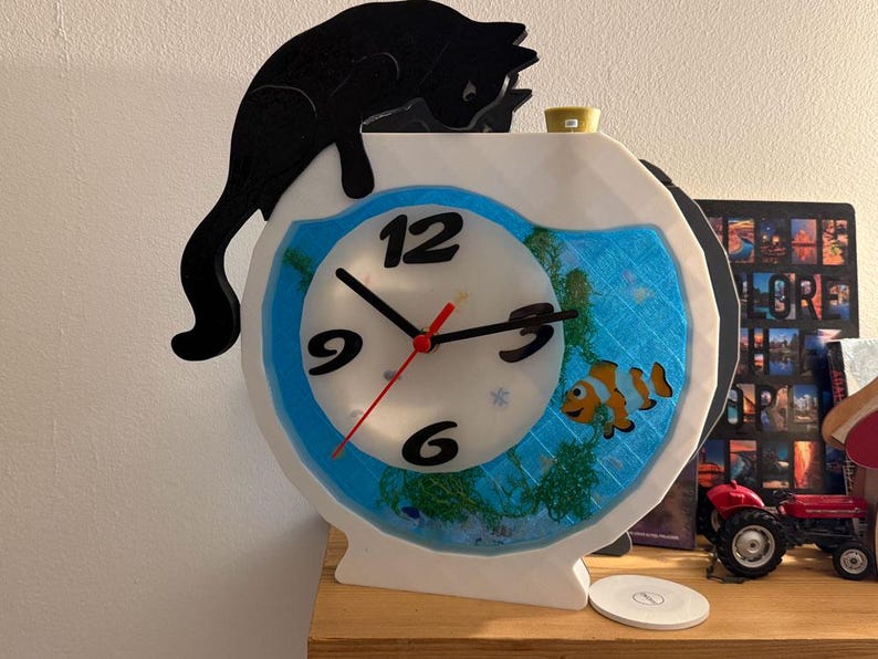 May include: A whimsical clock shaped like a fishbowl, featuring a clownfish and aquatic plants. A black cat silhouette perches atop the white frame. The clock face has black numbers and hands, with a red second hand. A unique decorative item.