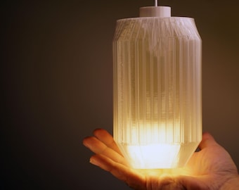 May include: A white, cylindrical pendant light fixture with vertical grooves. The light is illuminated from within, casting a warm glow. The fixture is held in a hand against a dark background.