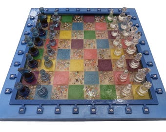 May include: A colorful, handcrafted chess set with a blue wooden frame. The board features a mosaic of squares in various colors, with clear glass chess pieces. The pieces are arranged for a game in progress.