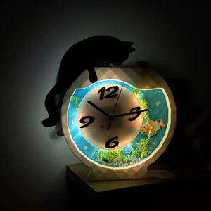 May include: A decorative clock with a fish tank design. The clock face features a blue background with a fish and aquatic plants. A black cat silhouette sits atop the clock. The clock is illuminated with a soft light.