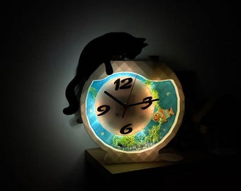 May include: A decorative clock with a fish tank design. The clock face features a blue background with a fish and aquatic plants. A black cat silhouette sits atop the clock. The clock is illuminated with a soft light.