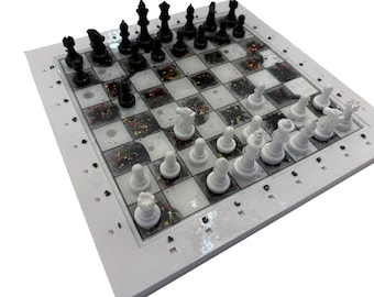 May include: A complete chess set with black and white pieces arranged on a square board. The board has a white frame and a checkerboard pattern with clear squares filled with colorful glitter.