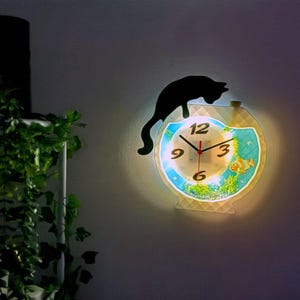 May include: A decorative wall clock featuring a black cat silhouette perched above a fishbowl design. The clock face has a light blue background with fish and plant illustrations. The clock hands are red.