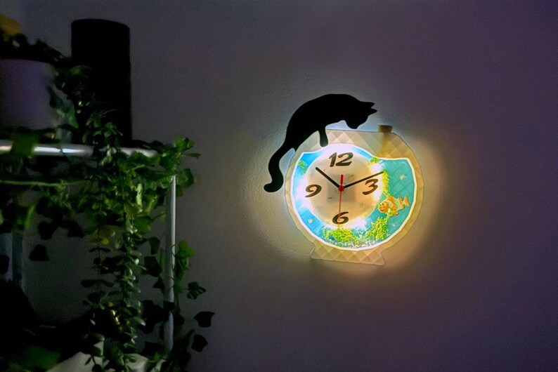 May include: A decorative wall clock featuring a black cat silhouette perched above a fishbowl design. The clock face has a light blue background with fish and plant illustrations. The clock hands are red.