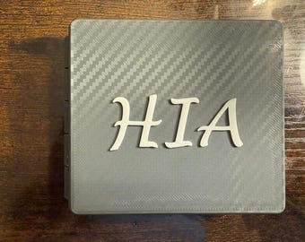 May include: A gray rectangular object with a carbon fiber pattern and the white letters "HIA". The letters are in a stylized font. The object appears to be a wallet or a similar item.