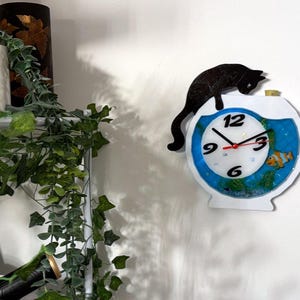 May include: A whimsical wall clock shaped like a fishbowl, featuring a black cat silhouette perched on top. The clock face has a blue border, black numerals, and red hands. A decorative fish and aquatic plants are visible inside the clock.