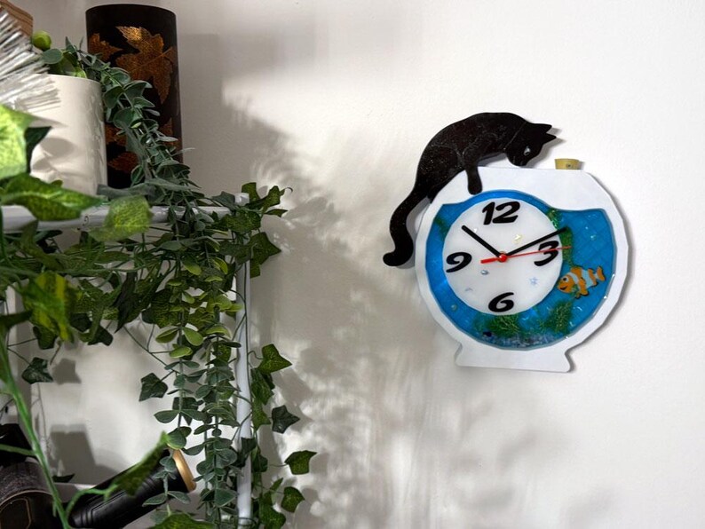 May include: A whimsical wall clock shaped like a fishbowl, featuring a black cat silhouette perched on top. The clock face has a blue border, black numerals, and red hands. A decorative fish and aquatic plants are visible inside the clock.