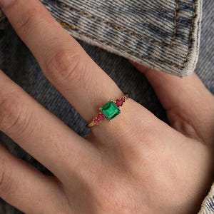May include: A gold ring featuring a square emerald-cut green gemstone as the center stone, flanked by two smaller, round, pink gemstones. The ring is set on a delicate gold band.