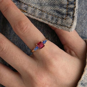 May include: A gold ring with a central rectangular, deep red gemstone and two smaller blue gemstones. The ring is displayed on a hand, with the jewellery highlighted against a denim jacket background.
