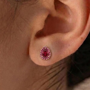 May include: A close-up of a person's ear with a teardrop-shaped stud earring. The earring features a vibrant red center stone surrounded by a halo of smaller, matching stones. The setting appears to be a light gold color, creating a striking contrast.