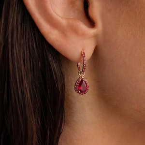 Könnte beinhalten: Nahaufnahme eines goldfarbenen Creolen-Ohrrings mit einem tropfenförmigen roten Edelsteinanhänger. Der Reifen ist mit kleinen roten Edelsteinen besetzt. Der Ohrring wird am Ohr einer Person getragen.