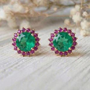 Puede incluir: Un par de pendientes de botón dorados. Cada pendiente presenta una piedra central grande, redonda, de color verde esmeralda, rodeada por un halo de piedras más pequeñas, redondas, de color magenta. Los pendientes están sobre un fondo borroso de flores blancas.