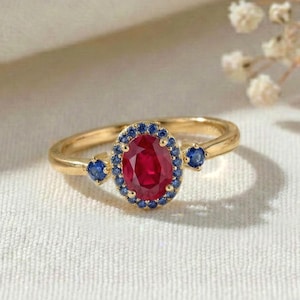 May include: A gold ring featuring a central oval-cut red ruby surrounded by a halo of small blue sapphires. Two additional blue sapphires are set on the band, one on each side of the central stone, creating a three-stone style with a halo design.