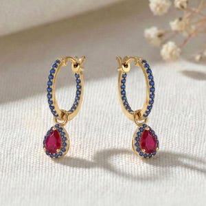 May include: A pair of gold hoop earrings featuring a circular band encrusted with small blue stones and a single teardrop-shaped red stone pendant hanging from the bottom of each hoop, creating a vibrant contrast of colors and shapes.