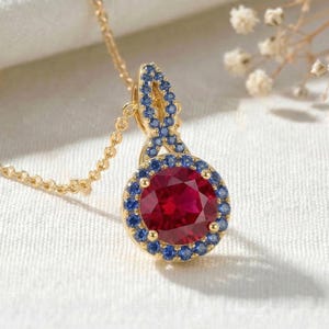 May include: A gold pendant necklace featuring a large, round, deep red centre stone surrounded by a halo of small blue stones. The pendant has an additional decorative loop at the top also set with small blue stones, suspended on a gold chain.