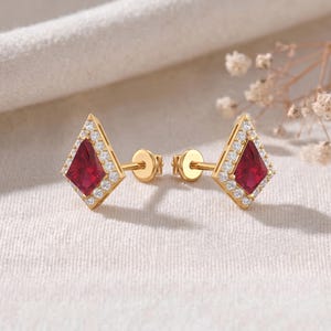 May include: A pair of kite-shaped stud earrings crafted from yellow gold. Each earring features a central deep red gemstone in a kite cut, surrounded by a halo of small, clear, round-cut white stones. The earrings include gold post backings.