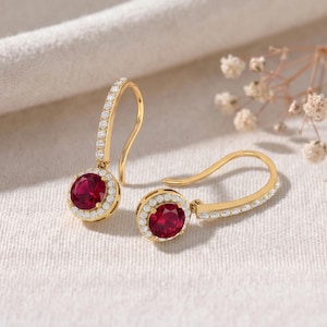 May include: A pair of gold drop earrings featuring round, deep red centre stones surrounded by a halo of small, clear white stones. The curved ear wires are also accented with a row of small, clear white stones along the front surface.