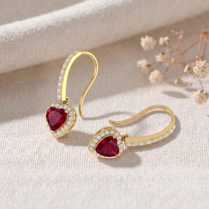 May include: A pair of gold drop earrings featuring heart-shaped red gemstones surrounded by small white stones. The curved ear wires are also accented with a row of tiny white stones, creating a cohesive and elegant design for these jewellery pieces.