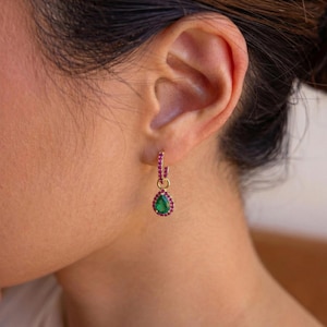 Puede incluir: Pendientes de aro dorados con un colgante en forma de lágrima. El aro está adornado con pequeñas piedras preciosas rosas, y el colgante presenta una gran piedra preciosa verde rodeada de piedras preciosas rosas más pequeñas. Los pendientes se llevan en la oreja de una persona.