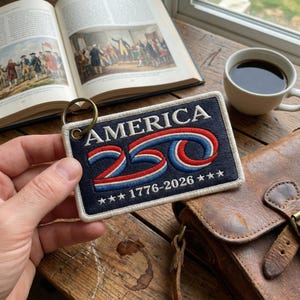 May include: A rectangular keychain patch with the word "AMERICA" in white, and "250" in red, white, and blue. The text "1776-2026" and three stars are below. The patch is on a wooden surface, with a cup of coffee and a book in the background.