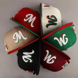 May include: A collection of baseball caps in various colors, including red, green, black, and tan. Each cap features a white script letter "M" or "W" with green and red accents. The caps have a flat brim and a small flag emblem.