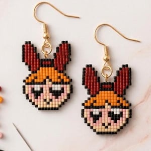 May include: A pair of beaded dangle earrings featuring a pixelated character design with orange hair, a red bow, and black outlines. The earrings are crafted from small square beads and hang from gold-toned metal hooks. A sewing needle and loose beads are nearby.