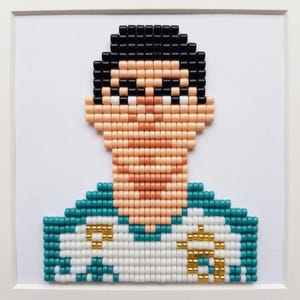 May include: A framed pixel art portrait created with small square beads. The figure has black hair, peach-colored skin, and wears a white shirt with teal accents and gold-colored details. The artwork is set against a plain white background.