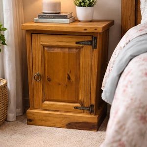 May include: A wooden nightstand with a single door and a small drawer. The nightstand is made of light brown wood and has a rustic design. A lamp, books, and a potted plant sit on top of the nightstand. The nightstand is next to a bed.