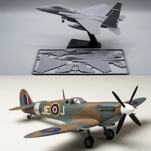 May include: A gray plastic model fighter jet on a black stand with an unassembled parts sprue, and a painted model propeller plane with brown and green camouflage, British roundels, and the identification code MJ143 JE-J on the side.