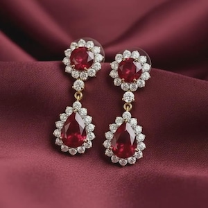 Puede incluir: Un par de elegantes pendientes colgantes con grandes piedras preciosas de color rojo intenso. Cada pendiente tiene una parte superior redonda en forma de flor con una piedra roja central rodeada de piedras claras más pequeñas.