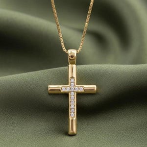 May include: A gold cross pendant necklace with a fine chain. The cross is embellished with small, sparkling gemstones. The necklace is set against a green backdrop, accentuating the jewellery's details.