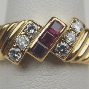 May include: A gold ring with a ribbed band design. The ring features three clear diamonds on each side of a rectangular setting with three red gemstones. The ring is set on a white surface.