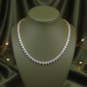 May include: A gold-toned necklace with a series of heart-shaped, clear gemstones. The necklace is displayed on a green jewelry stand against a backdrop of green fabric. The gemstones are evenly spaced along the necklace's chain.