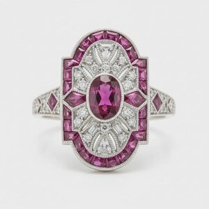 Puede incluir: Anillo de plata con una piedra central ovalada de color magenta, rodeada de piedras más pequeñas de color magenta y blanco. El anillo presenta un diseño Art Déco con formas geométricas y detalles intrincados.
