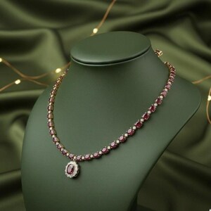 Puede incluir: Un collar dorado con piedras preciosas rojas de corte ovalado y pequeñas piedras blancas. El collar se exhibe en un busto de joyería verde. El colgante presenta una gran piedra roja ovalada rodeada de pequeñas piedras blancas.