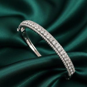May include: A silver bracelet with two rows of sparkling, clear gemstones. The bracelet is set against a backdrop of deep emerald green satin fabric, creating a luxurious and elegant aesthetic.