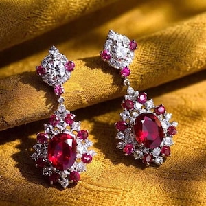 May include: A pair of drop earrings with a large oval ruby surrounded by smaller rubies and white gemstones. The earrings have a diamond-shaped top with a central stone and ruby accents, set in silver metal. The earrings are a luxurious piece of jewellery.