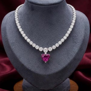 Puede incluir: Un collar de diamantes con un colgante de piedra preciosa rosa en forma de corazón. El collar presenta una serie de piedras preciosas redondas y claras engastadas en un metal plateado o blanco. El colgante es de color rosa vibrante.