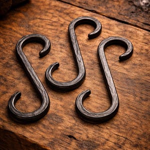 May include: Three handcrafted, dark metal S-hooks with a textured finish, arranged on a rustic wooden surface. The hooks have a classic design, perfect for hanging items. A hammer and chain are visible in the background.