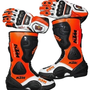 Könnte beinhalten: Ein Paar orangefarbene und schwarze KTM-Rennstiefel und passende Handschuhe. Die Stiefel und Handschuhe haben weiße Akzente und das Red Bull-Logo. Die Stiefel und Handschuhe sind für Motorradrennen konzipiert.