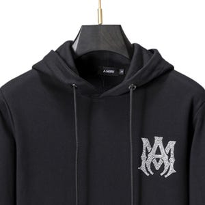 May include: Black hoodie with a drawstring hood and a large, ornate logo on the front. The logo is a stylized monogram in a lighter color, possibly silver or white. The brand name "AMIRI" is printed on the hood.