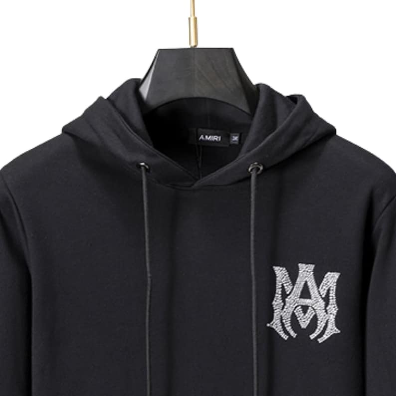 May include: Black hoodie with a drawstring hood and a large, ornate logo on the front. The logo is a stylized monogram in a lighter color, possibly silver or white. The brand name "AMIRI" is printed on the hood.