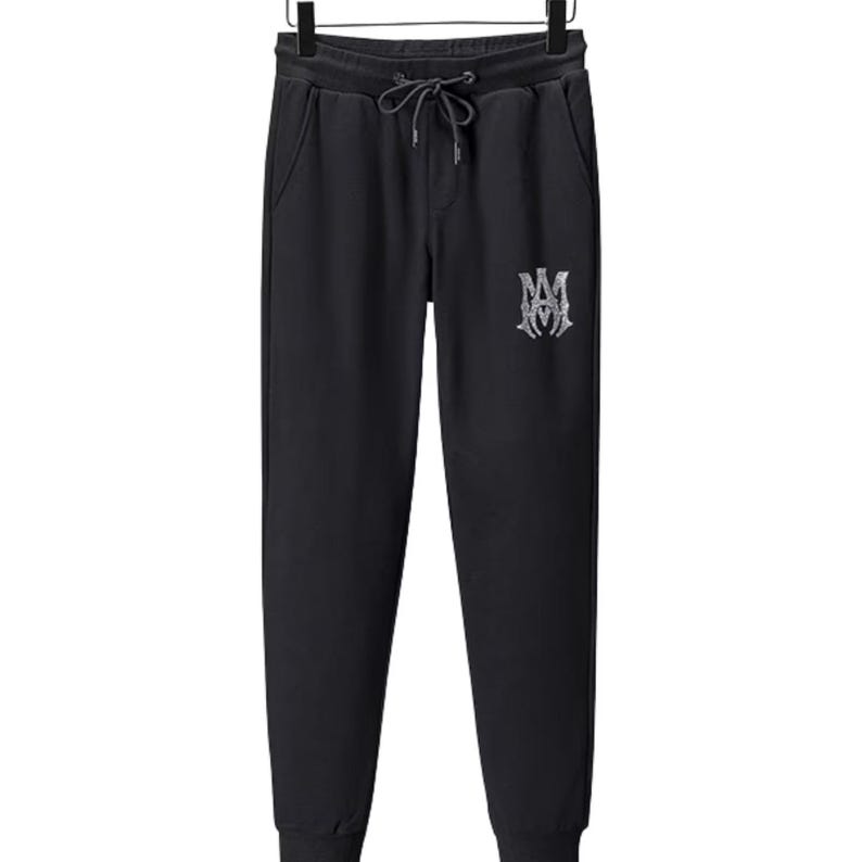 May include: Black sweatpants with a drawstring waist and side pockets. The pants feature a silver-colored embroidered logo on the left leg. The cuffs are ribbed. The pants are hanging on a black hanger.