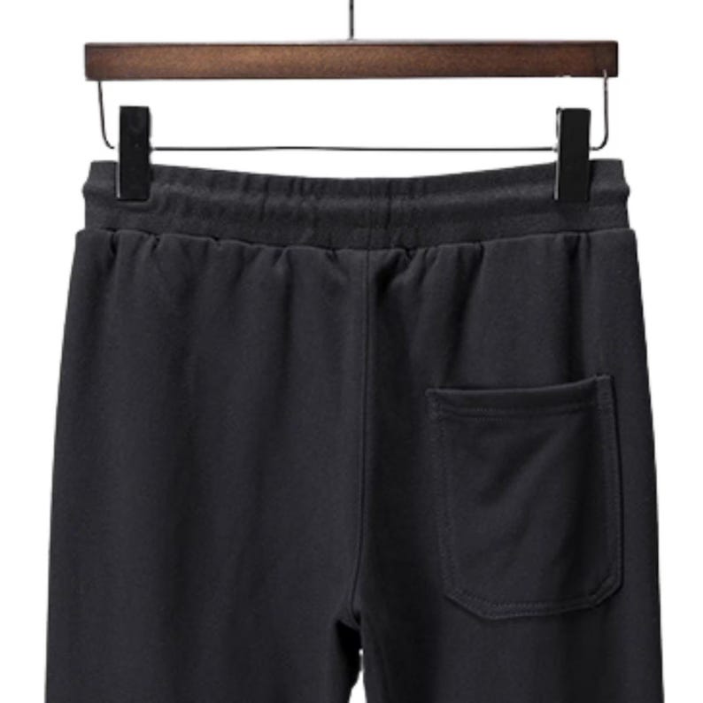 May include: Black sweatpants with a drawstring waistband and a single back pocket. The pants are hanging on a wooden hanger with a black metal hook. The fabric appears to be a soft, comfortable material.