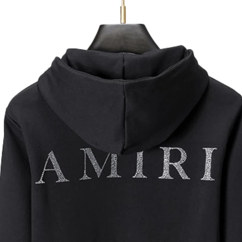 May include: Black hooded sweatshirt with the word "AMIRI" in silver, centered on the back. The hoodie has a drawstring and a hood. The fabric appears to be a soft material.